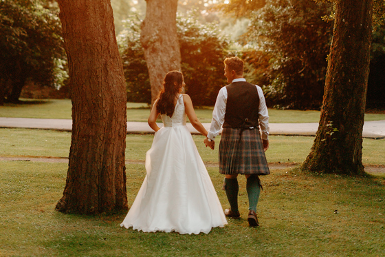 Bride and groom walking amongst trees at sunset holding hands